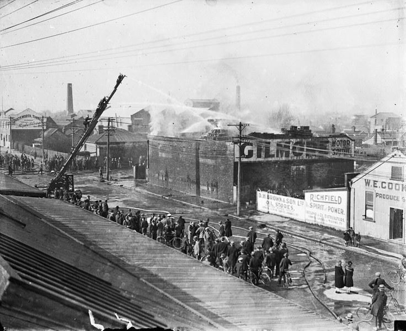 Fire at D.H. Brown Petrol Store, 1929. Gilmore was a US Brand of Petrol. Crowd of onlookers. On the left can be seen Duckworth and Turners Shoe Factory. Carlyle Street, Christchurch, Canterbury, New Zealand.