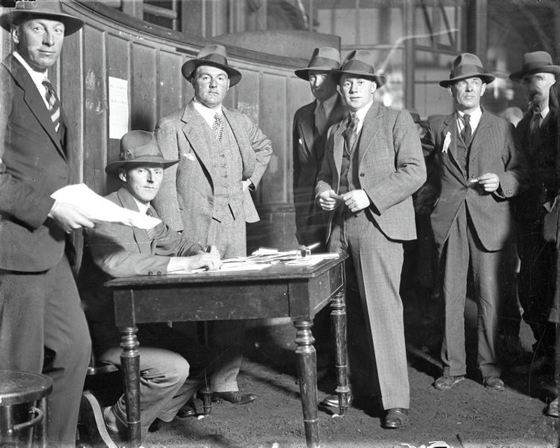 Men in a Panelled Room Line up at for a Man Seated at a Desk. All are in Suits, and are Wearing Hats. New Zealand.