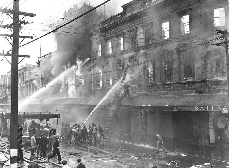 Ballantyne's Fire. Central City. Note Use of Fire Engine as a Pumping Engine. Firemen Struggle with Ladder on Balcony. Note use of Civilians to Help Manage Hoses. Also Note the Traffic Lights on the Pole in the Foreground, Colombo Street. Christchurch, Canterbury. New Zealand. 2