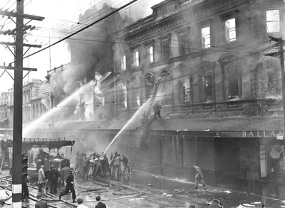 Ballantyne's Fire. Central City. Note Use of Fire Engine as a Pumping Engine. Firemen Struggle with Ladder on Balcony. Note use of Civilians to Help Manage Hoses. Also Note the Traffic Lights on the Pole in the Foreground, Colombo Street. Christchurch, Canterbury. New Zealand. 2 image item