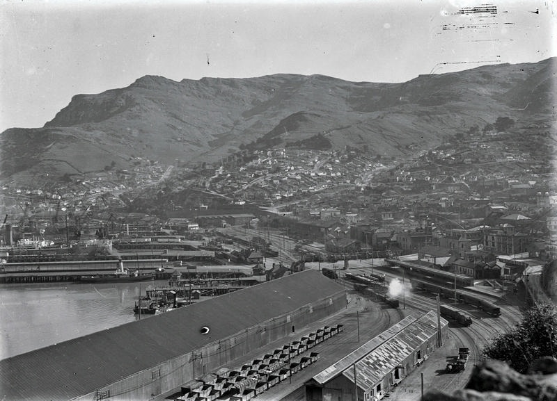View West across Lyttleton. On the Left at Wharf No2E, the Tug Lyttelton, 1908, 292 GRT, ( Gross Registered tons ) releases a wisp of smoke. On the Right of the Railway Yards Behind the Station can be seen the Roundhouse with its Turntable in Front. In the Foreground, Empty Railway Wagons Wait. At the Rear are Andersons Foundry and the Railway Tunnel Mouth. Lyttelton, Canterbury, New Zealand.
