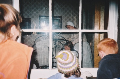 Visitors looking through a window at a Puhinui bedroom scene during a Candle lit tour at Howick Historical Village, 27 May 2003 showing a 'mother tending her daughters'. ; 27 May 2003; P2022.08.15
