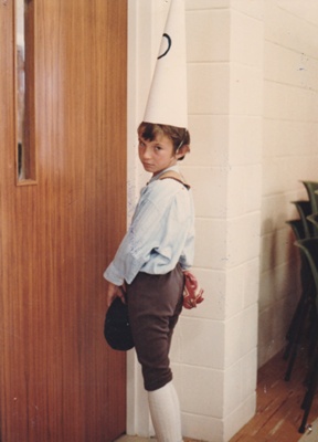 A boy in a dunce's hat standing in the corner in Pakuranga School in Howick Historical Village during a school visit.; N.Z. Woman's Weekly; 1982; P2021.182.05