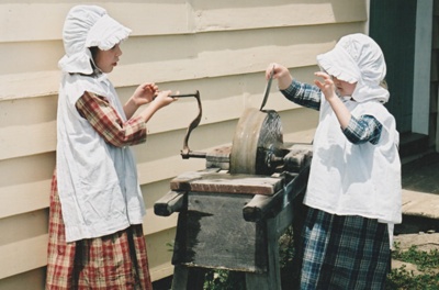 The two little girls in costume from the previous photos using the grindstone in Howick Historical Village. ; La Roche, Alan; P2020.18.18