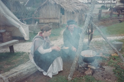 A man and woman in costume by the fireplace outside the tent at Howick Historical Village.; La Roche, Alan; P2021.88.20