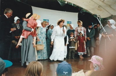 The November 1977 150th re-enactment of the Fencible and early settler landing at Cockle Bay. Shows Debbie Benson, extreme right behind microphone and other 'settlers' being welcomed to Howick by the lady in the white dress?. Sir Barry Curtis is extreme left on the stage.; November 1977; P2021.92.13
