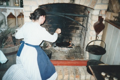 A woman, in costume, cooking in the fireplace in a cottage in Howick Historical Village.; La Roche, Alan; P2021.98.04