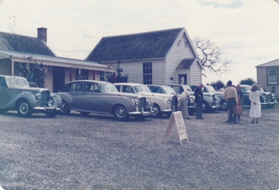 A line up of cars during the visit of the Rolls Royce-Bentley Car club to Howick Historical Village. Brindle Cottage and the Courthouse are behind.; La Roche, Alan; August 1985; P2021.108.19