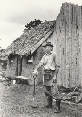 Arthur White holding a spade in front of the Sod Cottage, Howick Historical Village 
; 6 November 1980; P2020.43.17