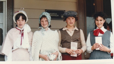 Three girls and a boy in costume, on a Gala weekend at Howick Historical Village, standing on the balcony at Eckford's Homestead, formerly Maraetai Cottage.; 1985; P2021.178.01