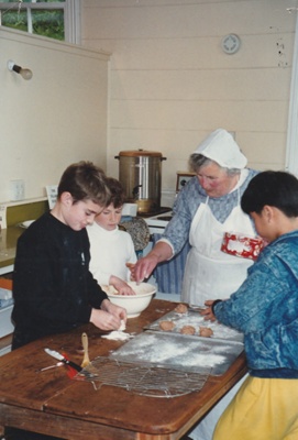 Three boys helping a Village guide with baking in Pakuranga School in the Howick Historical Village.; P2020.64.08