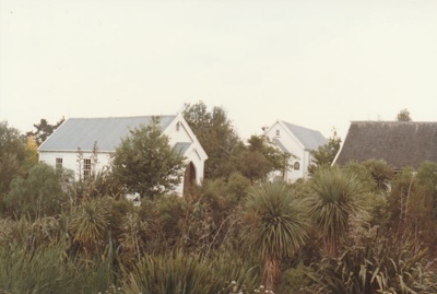 Looking across thick vegetation to the Howick Methodist Church, Ararimu Valley School and the roof of Sergeant Barry's cottage in the Howick Historical Village.; Ashby, J; 1984; P2020.34.13
