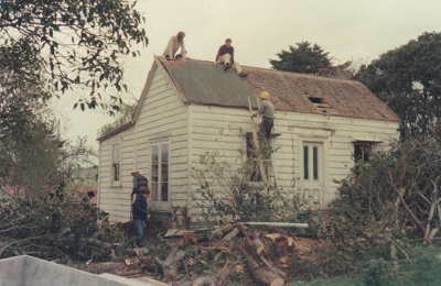 Kay Langdon's cottage at 33 Drake Street, Howick before its removal to Howick Historical Village to become Brindle Cottage. Shows men removing the shingles which were underneath an iron roof.; La Roche, Alan; June 1977; P2021.38.07