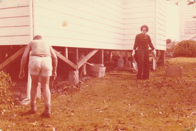 June Richardson standing beside McDermot's  cottage in Tainui Garden of Memories being prepared for removal to the Howick Historical Village.; La Roche, Alan; 12/05/1979; P2019.092.00