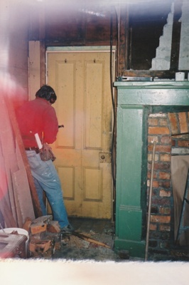 Puhinui kitchen during restoration, showing part of the fireplace, a man working on a door and building tools and debris.; Alan La Roche; September 2003; P2020.14.23