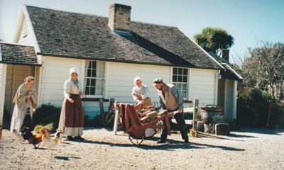 Richard Lees carting bricks outside  Briody-McDaniel Cottage. Prue Lees and two other women are also there. All are in costume. 
; La Roche, Alan; c2000; P2020.104.04