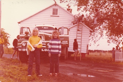 McDermott's Cottage on a transporter at Howick Historical Village jammed between an oak tree and a power pole. Warwick Johnson and David Mirams are about to prune the tree. Tania Marsh and Maria la Roche are standing in front.
; La Roche, Alan; 19 December 1974; P2020.100.13