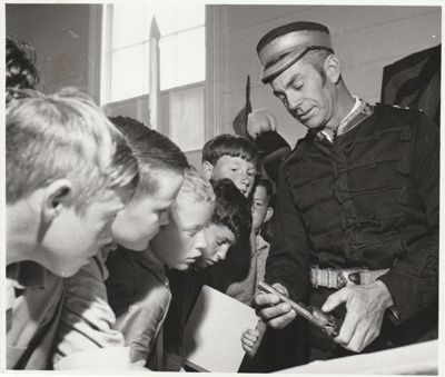 A 'soldier' demonstrating a pistol to a group of boys at the 1962 exhibition of the Howick Historical Society in the Howick Town Hall.; Auckland Star; 1962; P2022.10.10