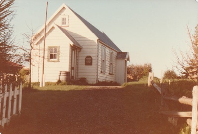 Ararimu Valley School in the Howick Historical Village with the addition of the side entrance porch. A flagpole and barrel are in front of the building. Also shows fences.; September 1984; P2020.20.08