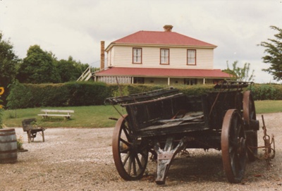 A wagon in state of disrepair at the Howick Historical Village. Bell House is in the background. A barrel, wheelbarrow and bench seat are also shown.; September 1980; P2020.18.14