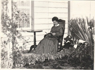 Eileen Martensen, Secretary of the Howick Historical Society reading while sitting in a chair outside a cottage at the 1962 exhibition of the Howick Historical Society in the Howick Town Hall.; Auckland Star; 1962; P2022.10.04