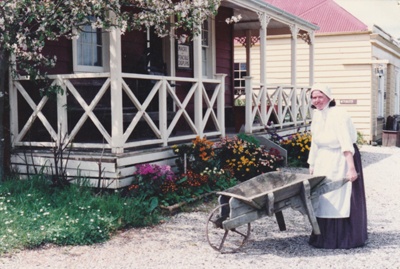 Debbie Benson in costume wheeling a barrow past Colonel de Quincey's Cottage on Grey Street at Howick Historical Village, with Johnson"s on the right.; P2020.108.03