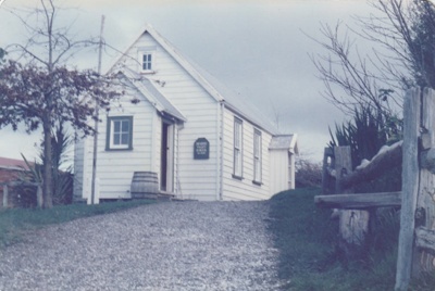 Ararimu Valley School in the Howick Historical Village with the addition of the side entrance porch. A flagpole and barrel are in front of the building. A fence is on the right side.; August 1986 or 1987; P2020.20.10