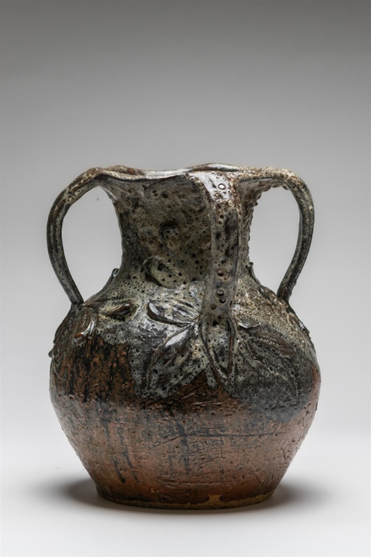 Around the base of the handles there are sculptured leaf motifs. The vase like vessel has a double glaze with light over dark drips and bubbles. Towards the bottom half of the vessel the overall effect is of a highly textured and scratched surface to the clay.