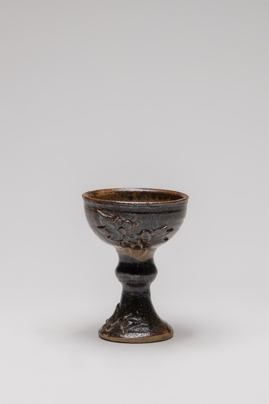 There is a dark glaze on the exterior of the goblet with no glaze in the interior of the cup. An unglazed strip starts from the top exterior of the cup, running down and along halfway around the base of the cup.