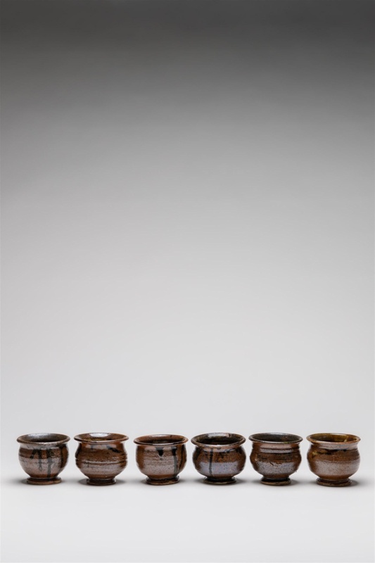 Set of six small squat cups. Dark glaze dripped over iron oxide glaze. Each cup is elevated with a small foot, emphasising the over-all shape.