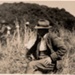 Photo, Man sitting in long grass, crowd in background; RAP2020.0067